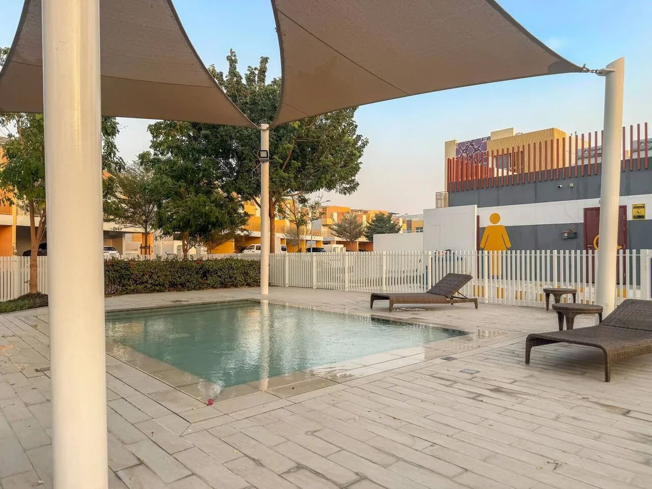 Una piscina al aire libre está bajo toldos de sombra, junto a una zona pavimentada con asientos.