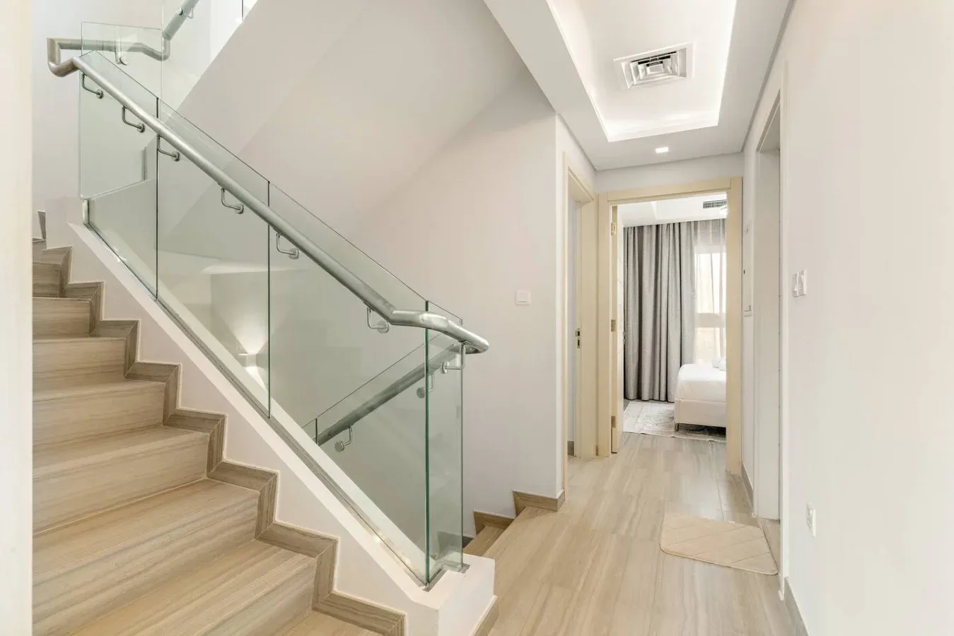 Un pasillo luminoso con una escalera y barandilla de vidrio conduce a un dormitorio.