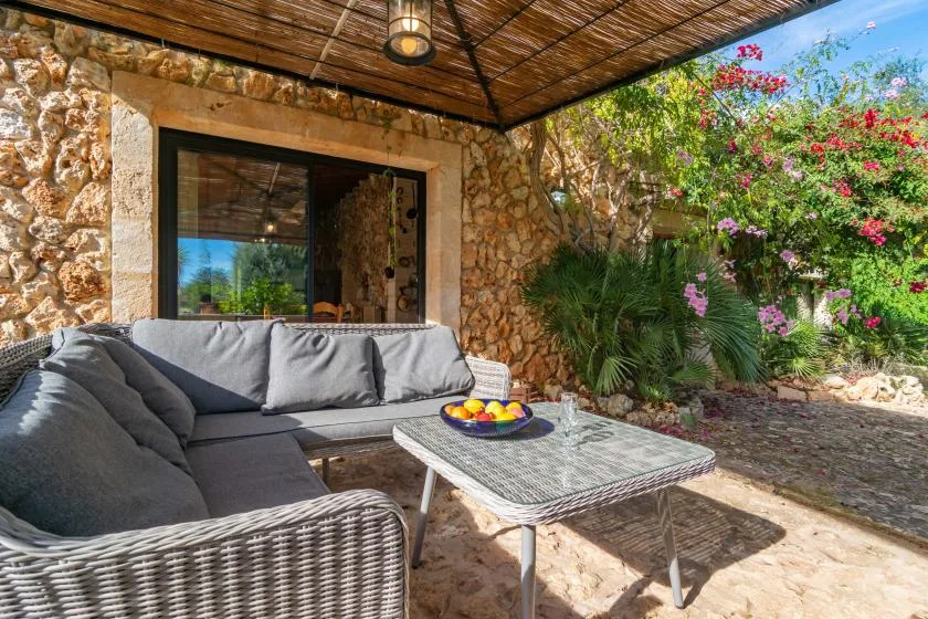 Una zona de estar exterior a la sombra, con una mesa junto a un muro de piedra y plantas con flores.