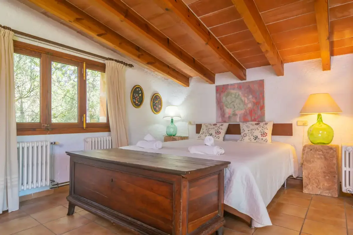 Un dormitorio con una cama grande, un baúl de madera y ventanas bajo un techo inclinado de vigas.