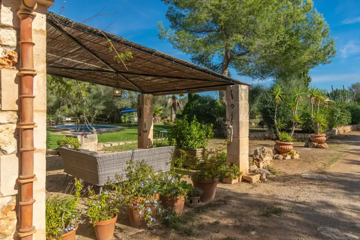 Un patio exterior sombreado con asientos y plantas en macetas da a un jardín.