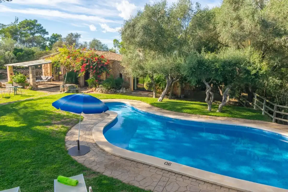 Una piscina curva atraviesa un jardín con césped, árboles y un patio sombreado.