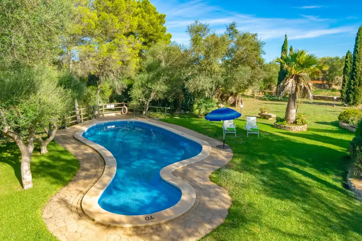 Una piscina sinuosa se encuentra en un jardín soleado rodeado de árboles.