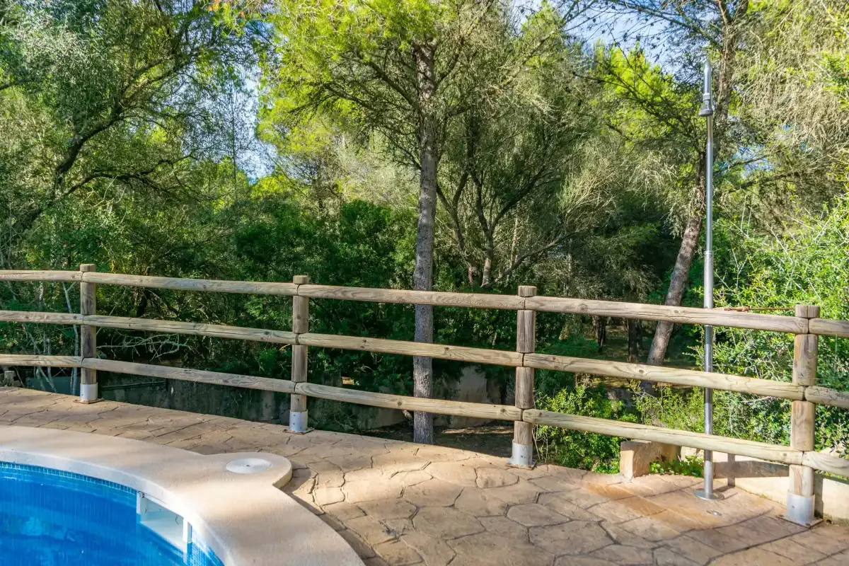Una piscina junto a un patio pavimentado y una barandilla de madera, rodeada de árboles.