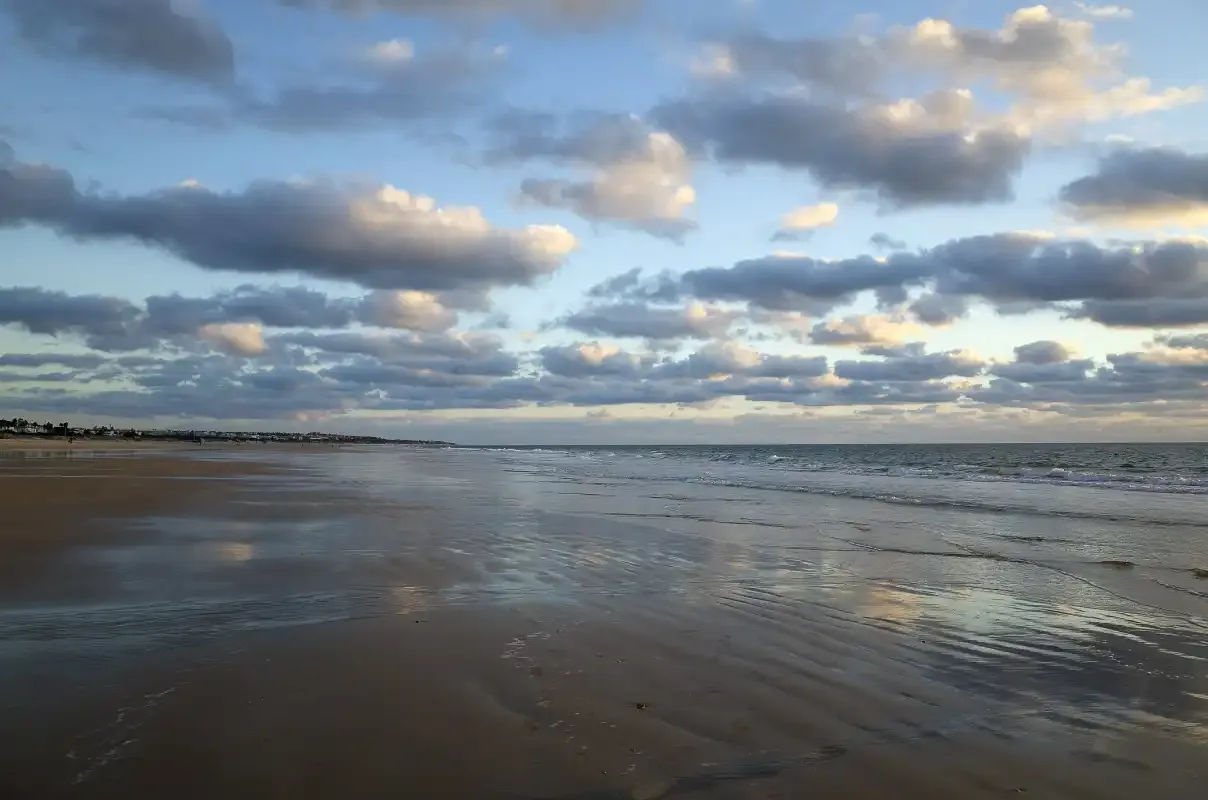 A broad beach at low tide reflects a cloudy sky.
