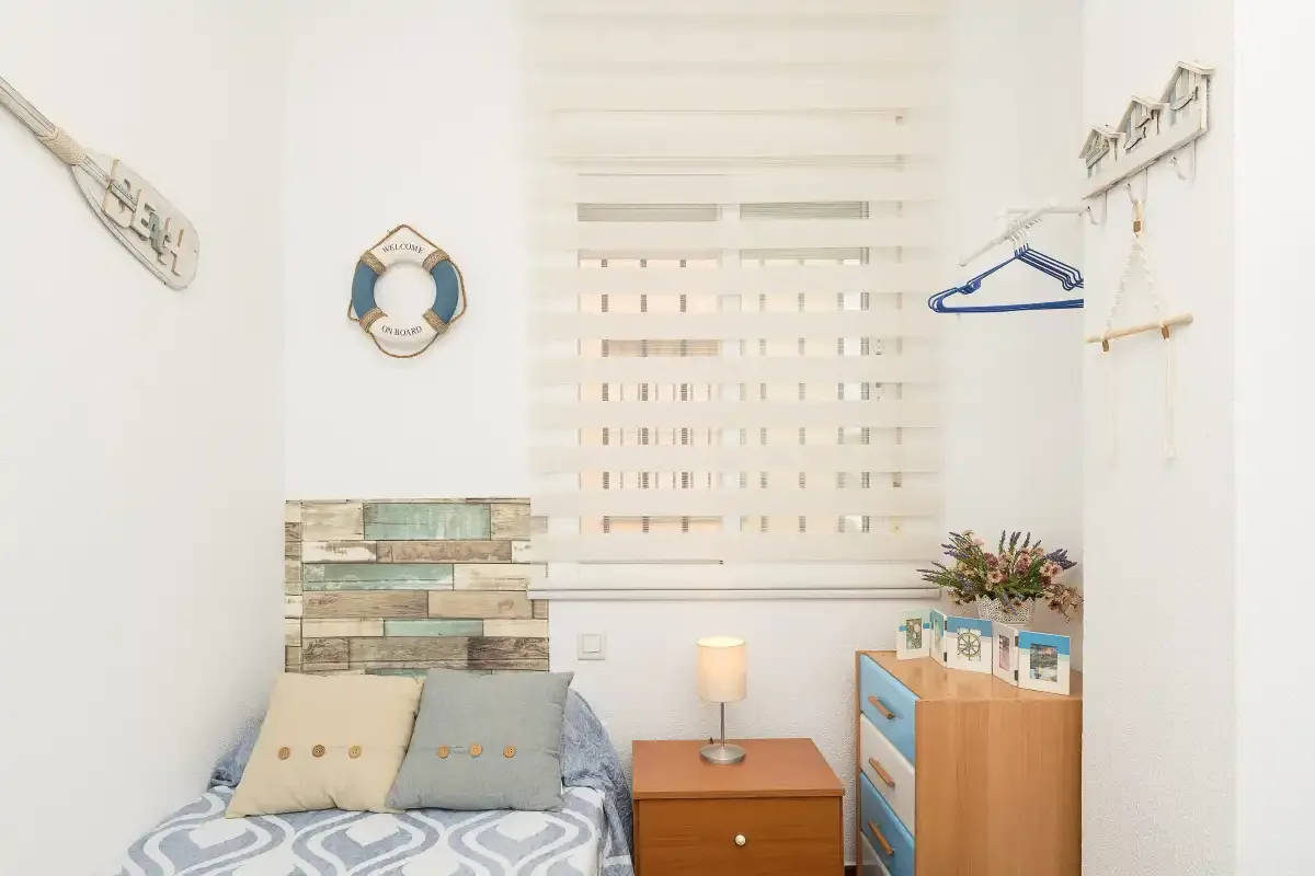 A bright bedroom with a bed, bedside table, and window blind.