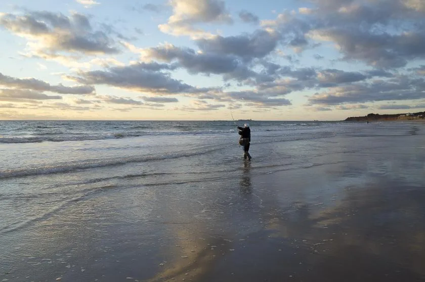 A person stands in shallow water on a beach beneath a cloudy sky.