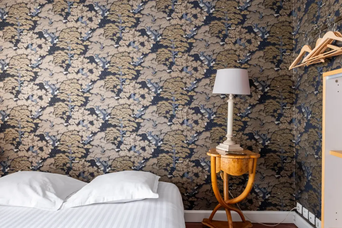 A bedroom with a bed, floral wallpaper, and a bedside table with a lamp.
