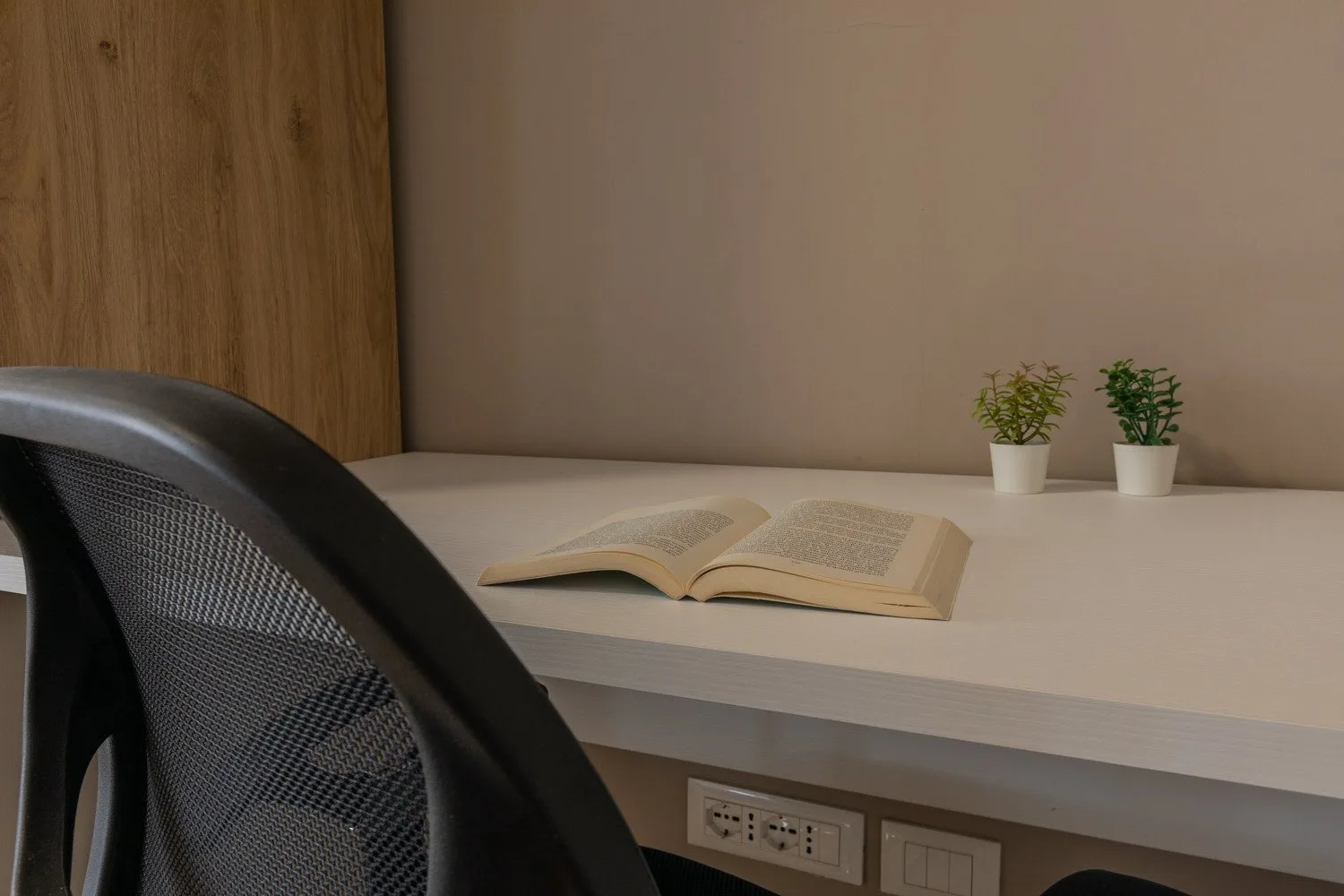 A desk with an open book and two small plants beside a mesh chair.
