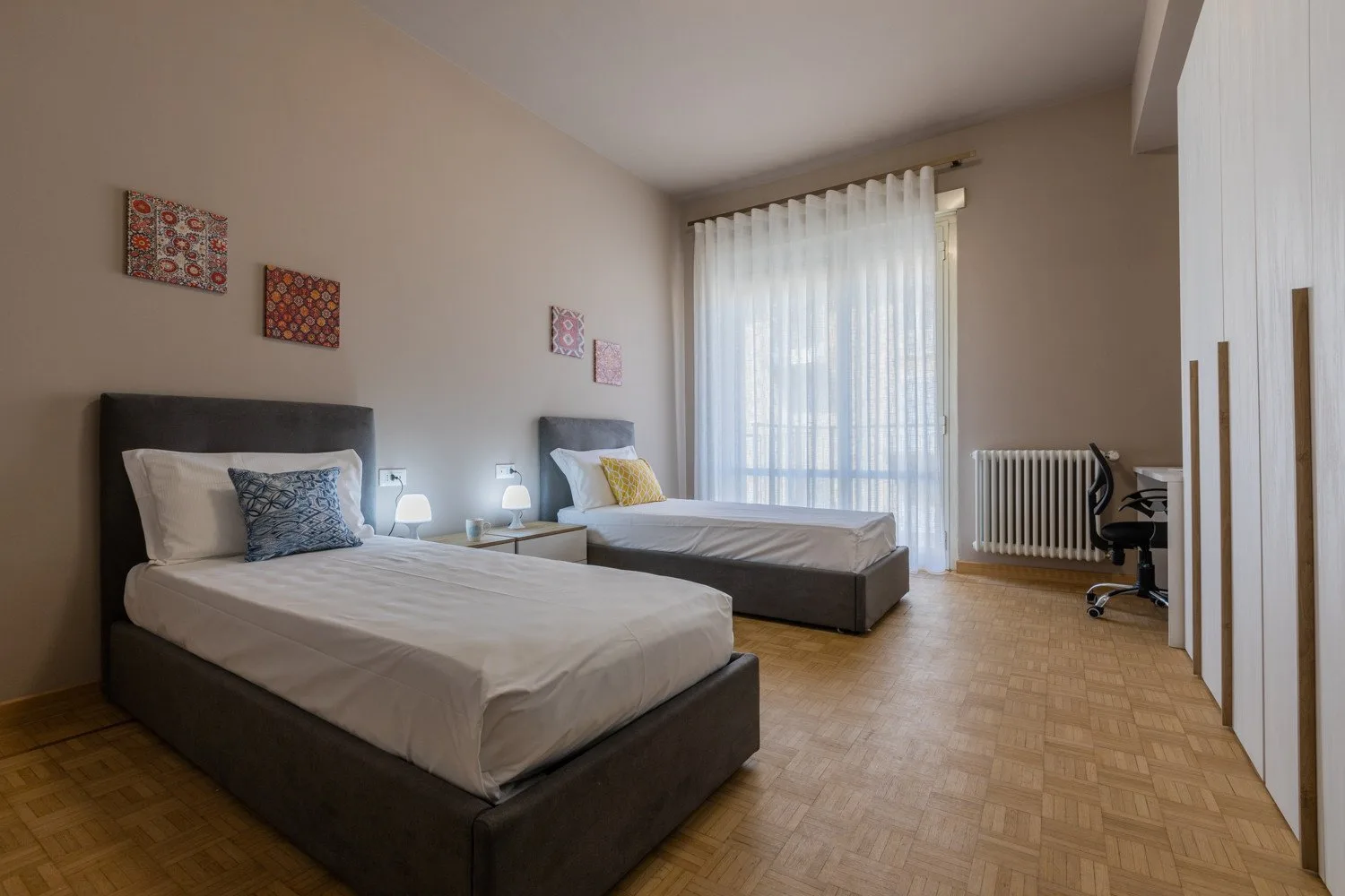 A bedroom with two beds, a desk, and a large curtained window.