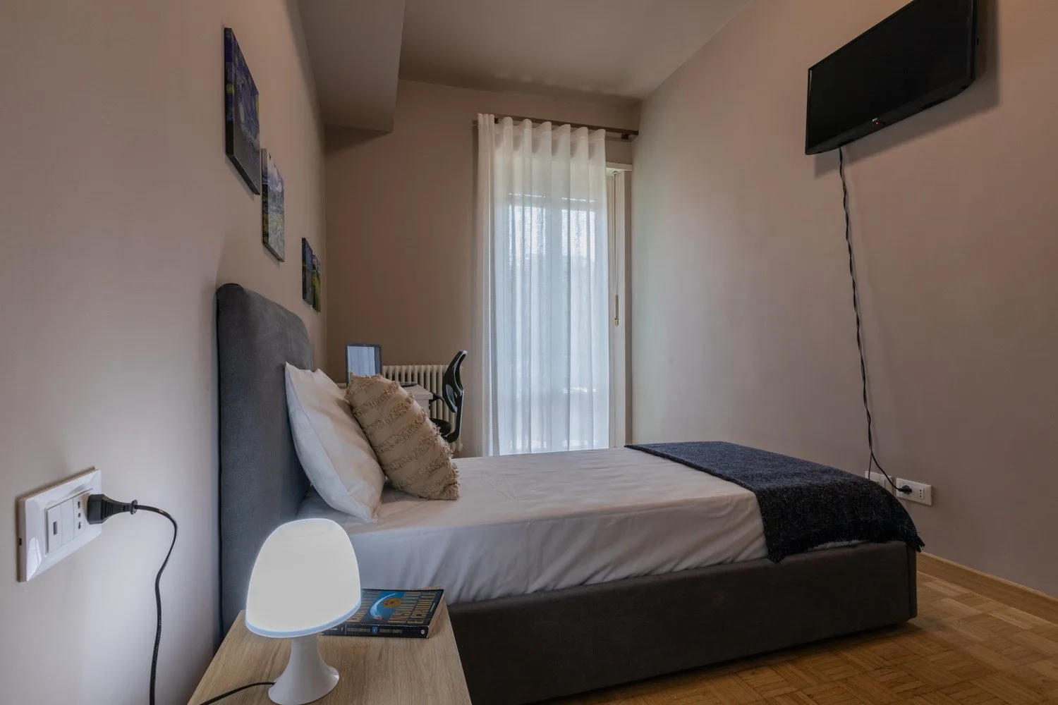 A bedroom with a bed beside a tall window with sheer curtains.