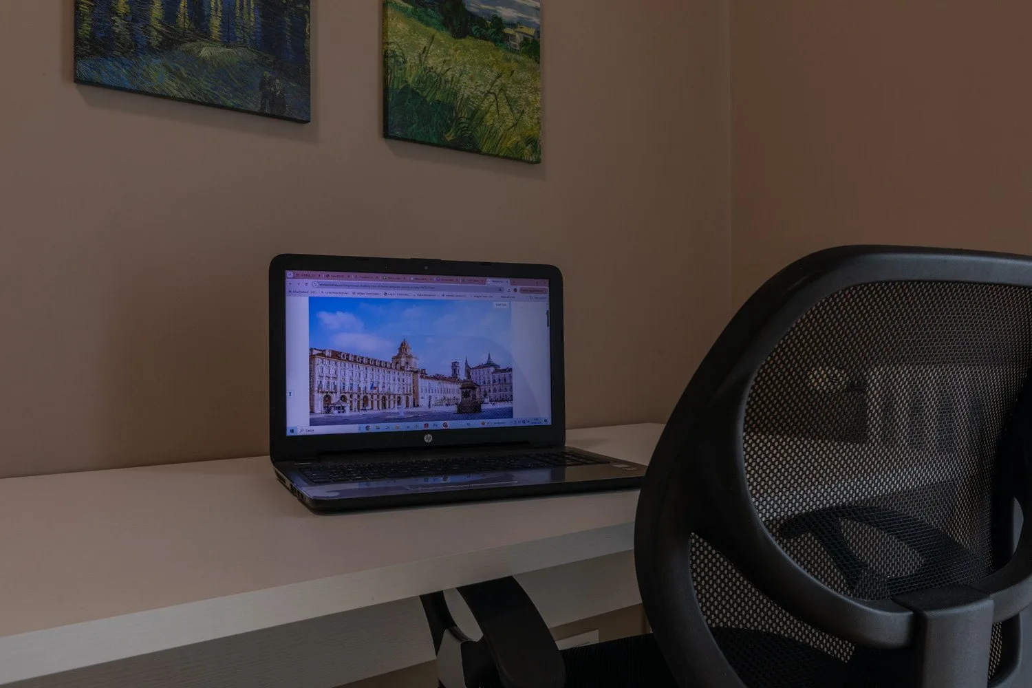 A desk with an open laptop and a mesh-backed chair sits beneath wall art.