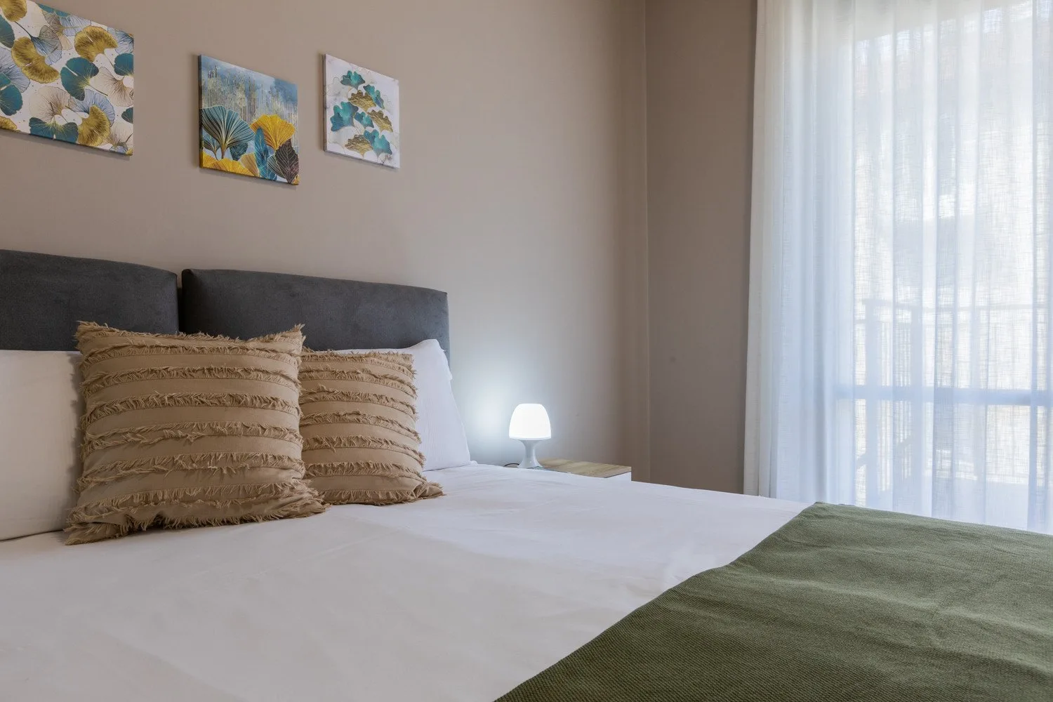 A bedroom with a neatly made bed and sheer curtains over a bright window.