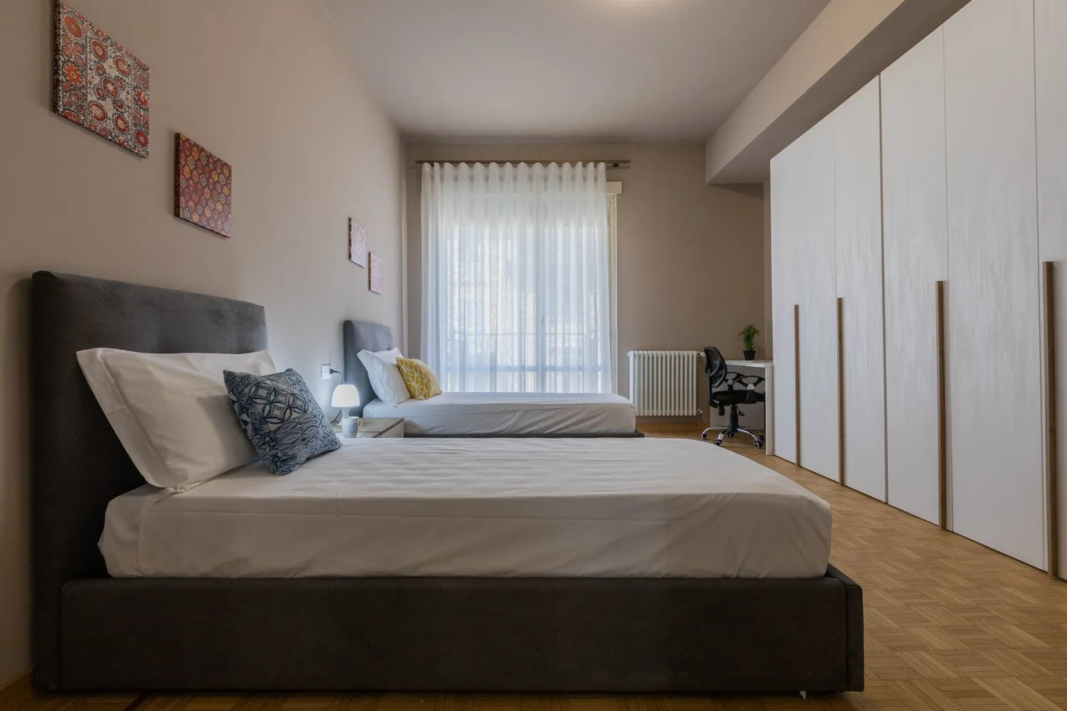A bedroom with two beds, a tall wardrobe, and a large curtained window.