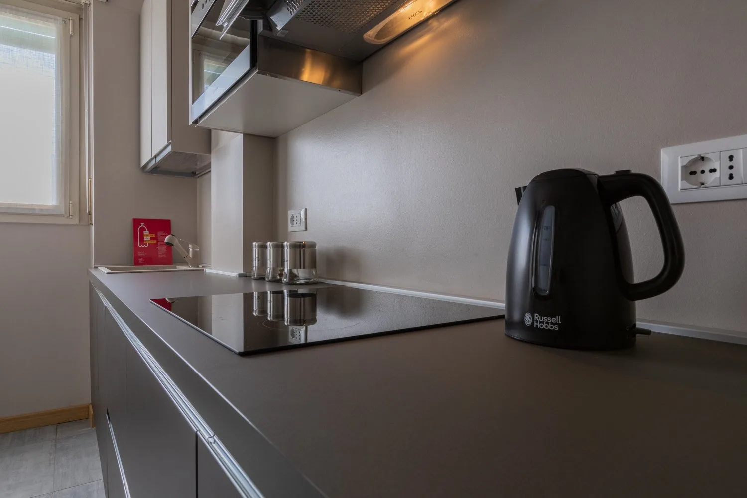 A compact kitchen counter with a kettle, cooktop, and overhead range hood.