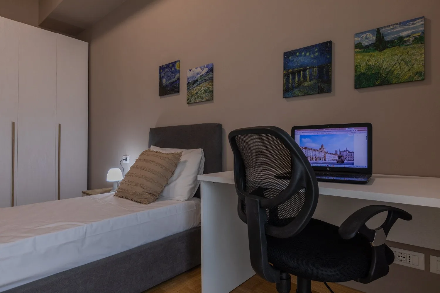 A bedroom with a bed, desk, and computer beneath several framed pictures.