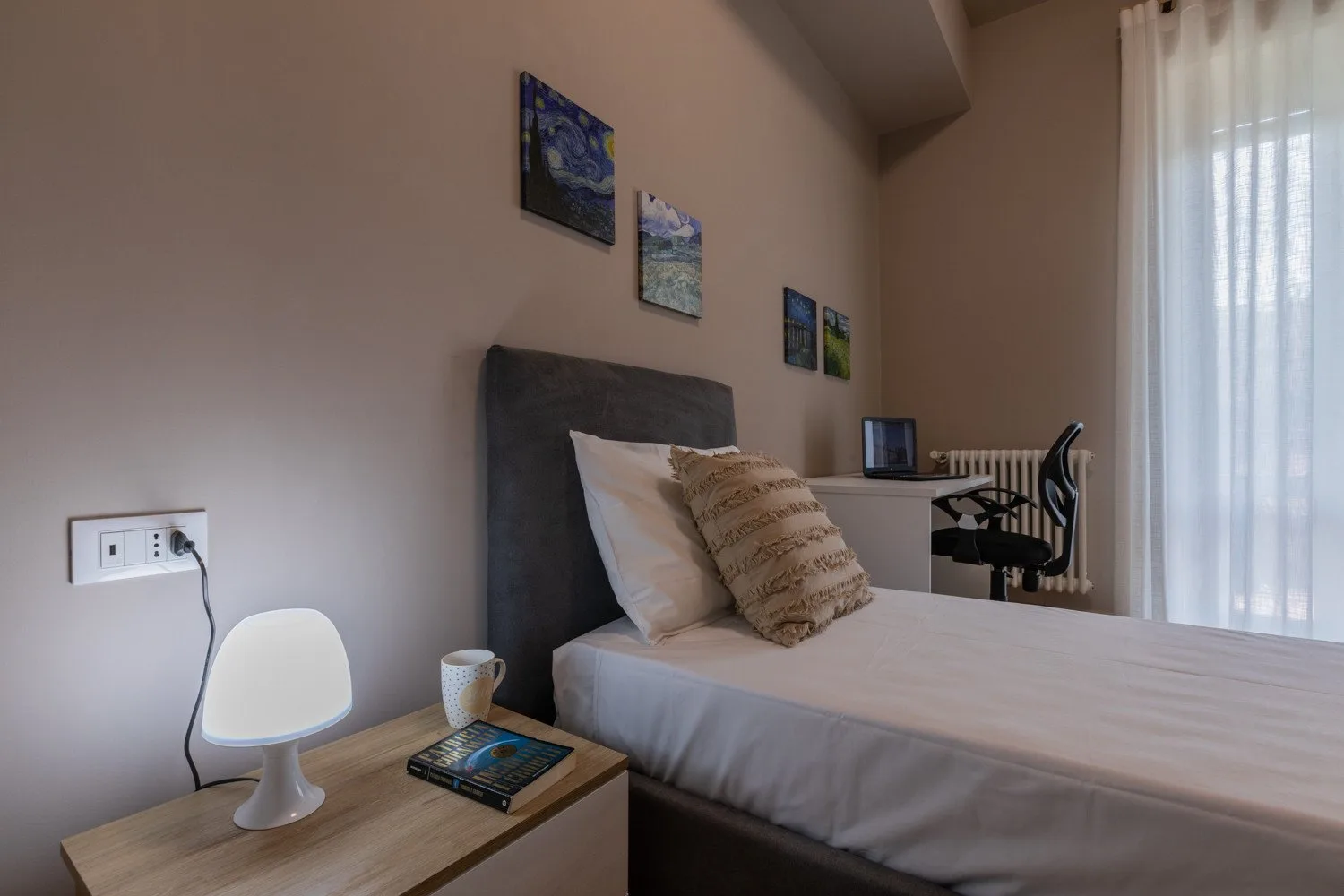 A bedroom with a bed beside a desk and a tall curtained window.