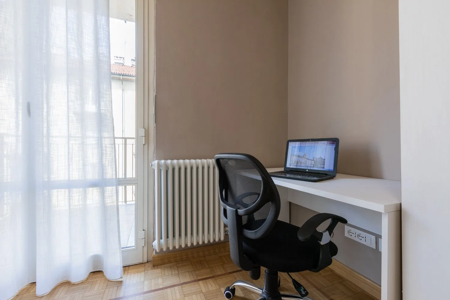 A desk with a computer and office chair sits beside a bright window.