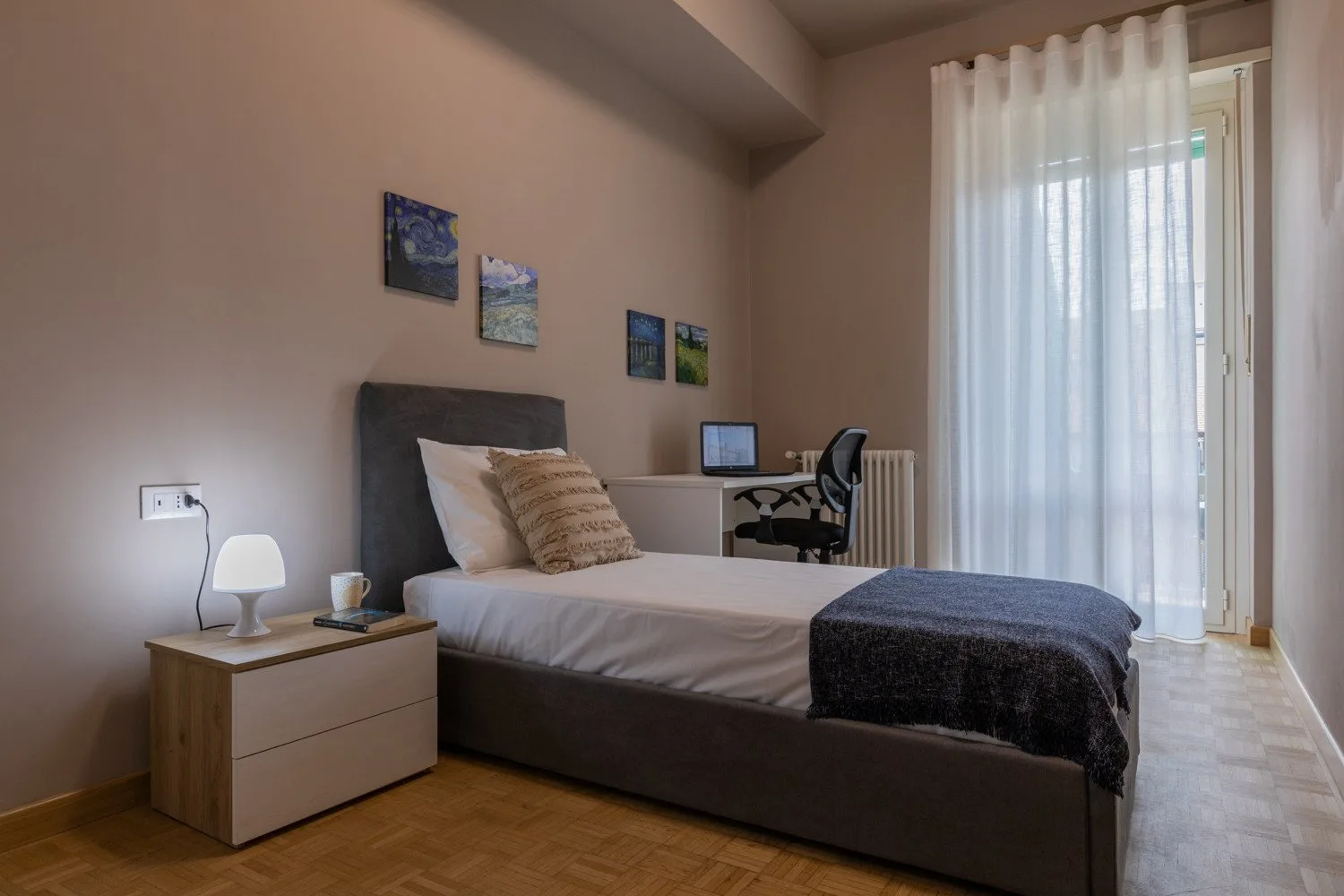 A bedroom with a bed, bedside table, and desk by a curtained window.