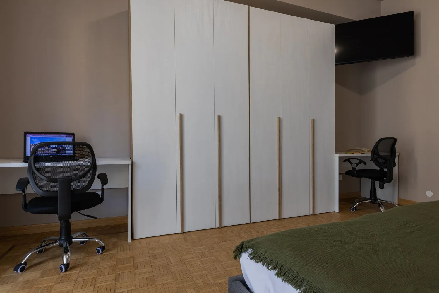 A bedroom with a large wardrobe, desk chairs, and a wall-mounted television.
