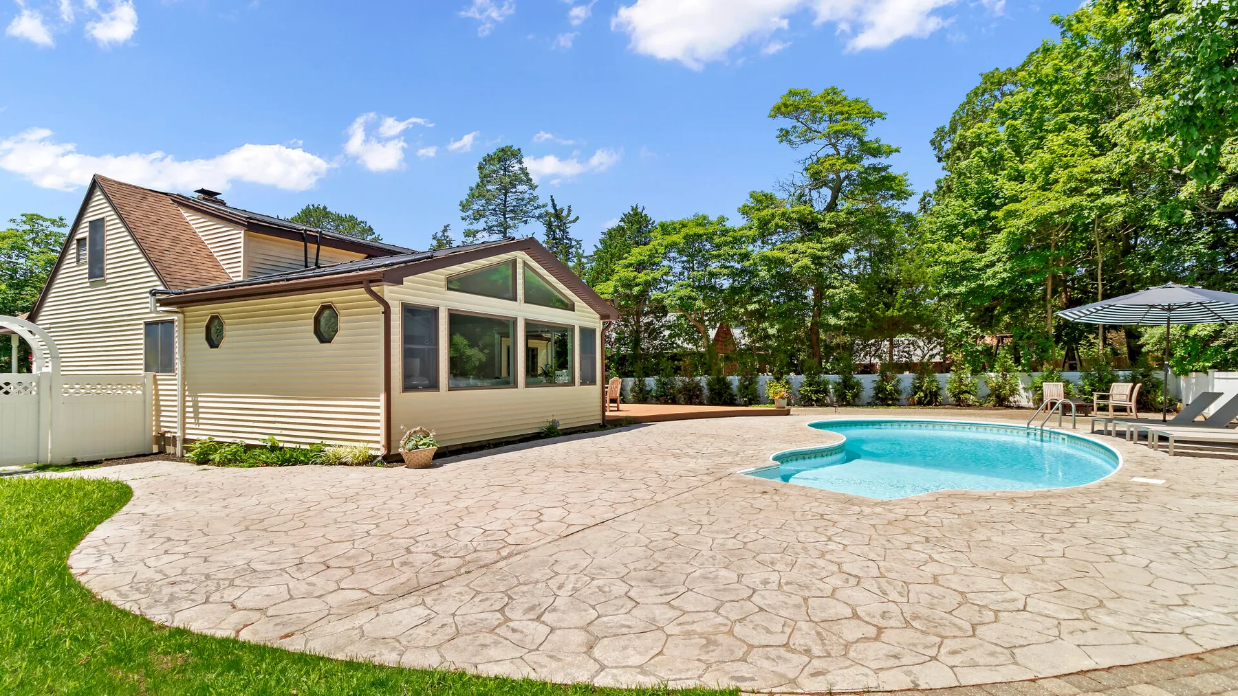 Una casa y una piscina están junto a un amplio patio pavimentado, rodeadas de árboles.