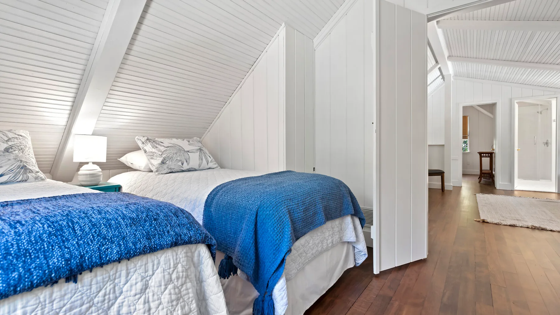 Un dormitorio abuhardillado luminoso tiene dos camas con ropa de cama azul bajo un techo inclinado.