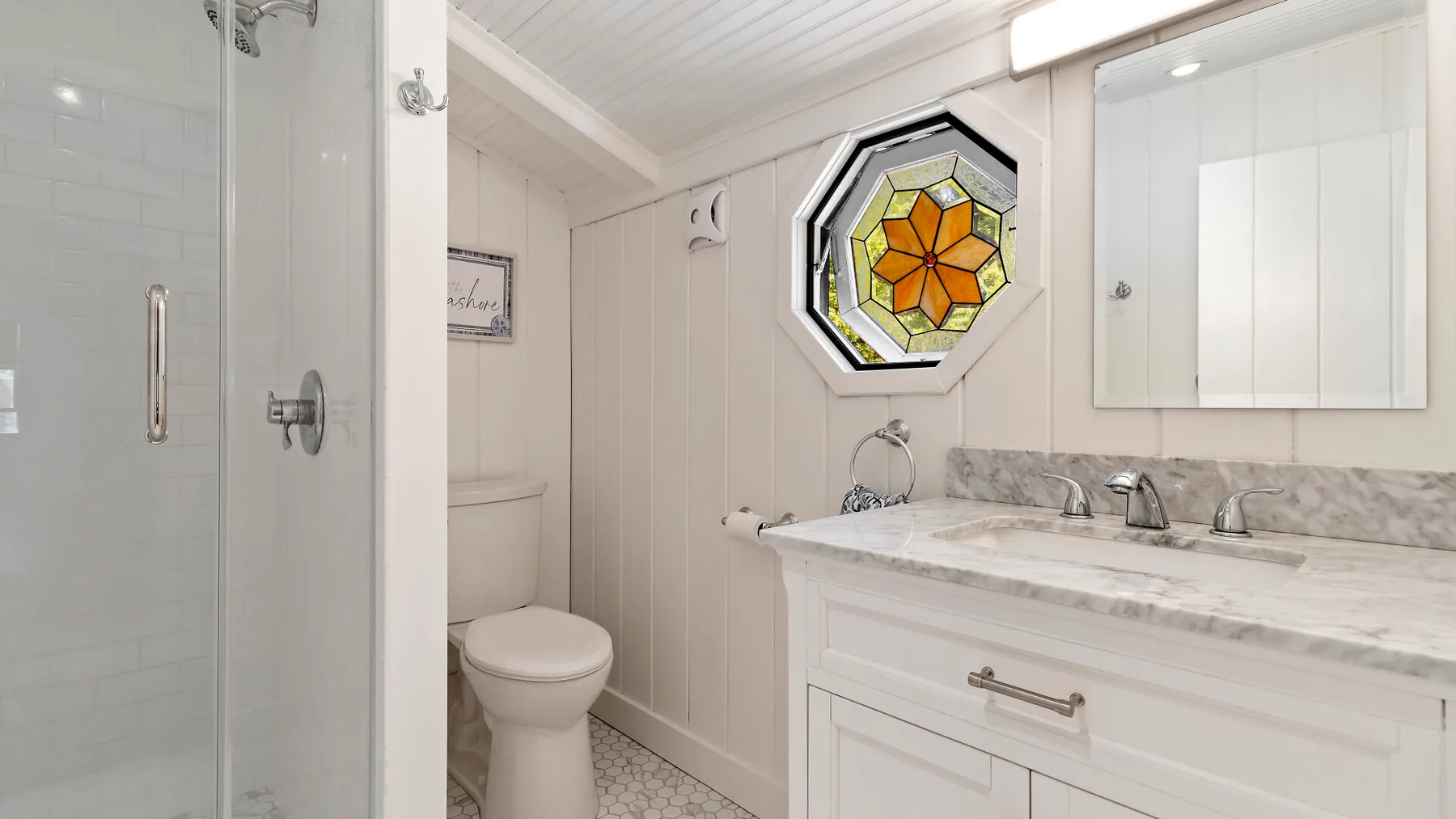 Un baño luminoso con tocador blanco, inodoro, ducha y una ventana de vidrio emplomado.