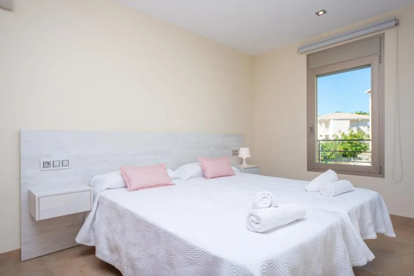 A bright bedroom with a white bed and a window overlooking nearby buildings.