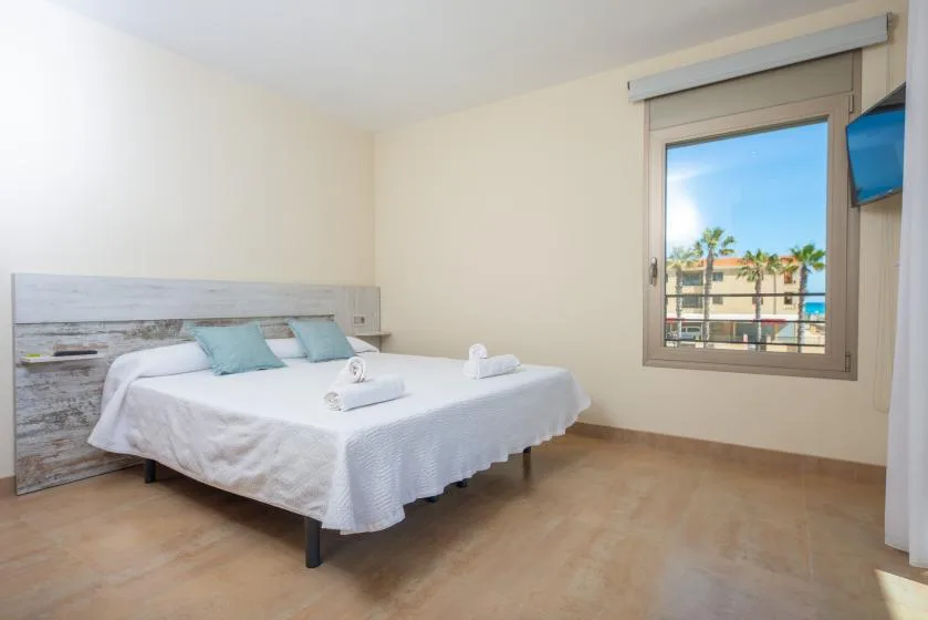A bright bedroom with white bedding and a window overlooking nearby buildings.