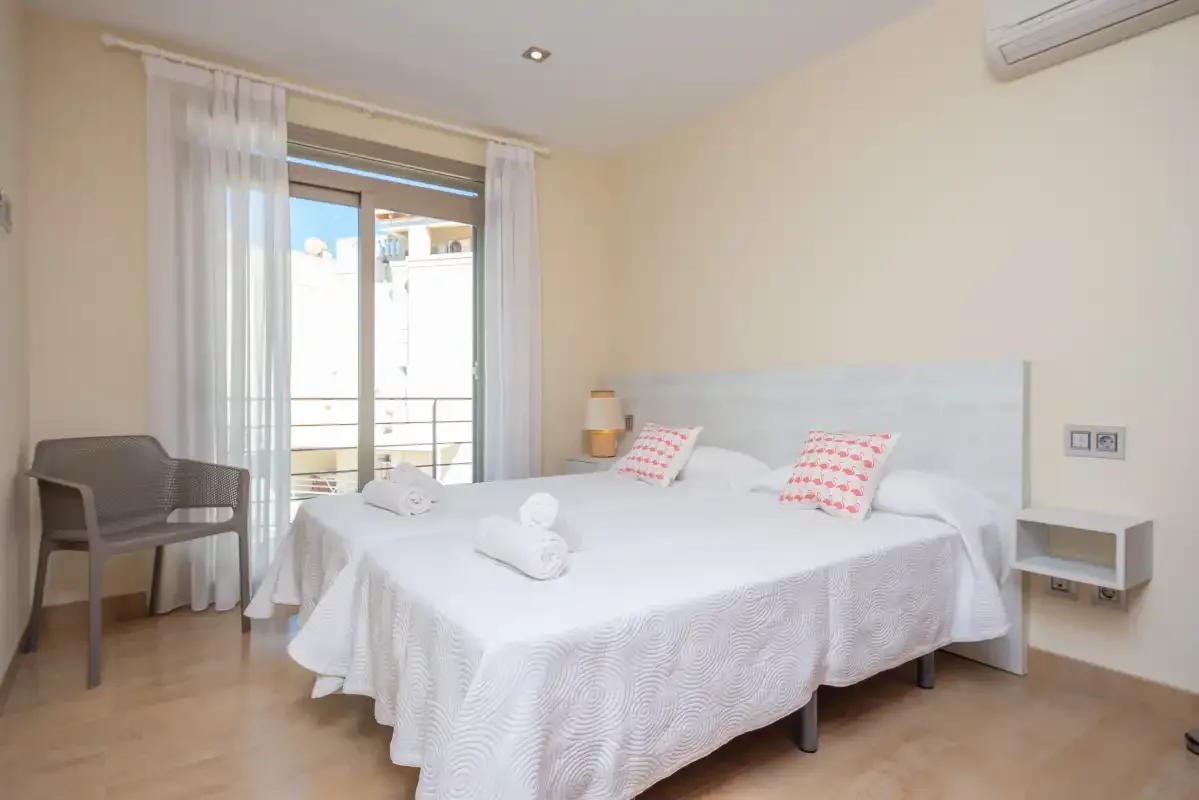 A bright bedroom with a neatly made bed beside a curtained balcony door.