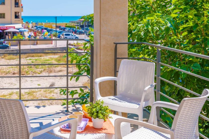 A balcony seating area overlooks nearby buildings, greenery, and the sea.