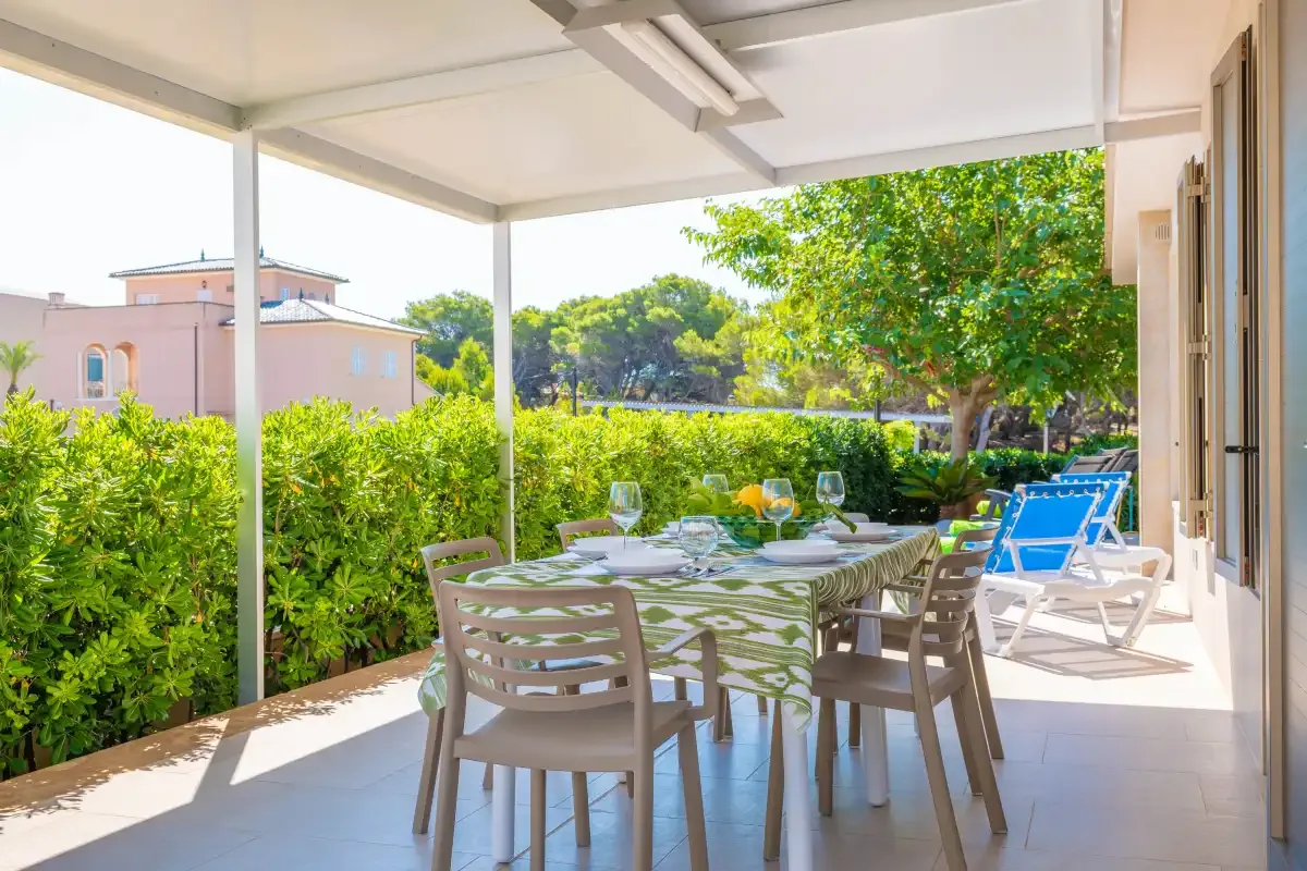 A covered outdoor dining area with a table and seating beside a leafy garden.