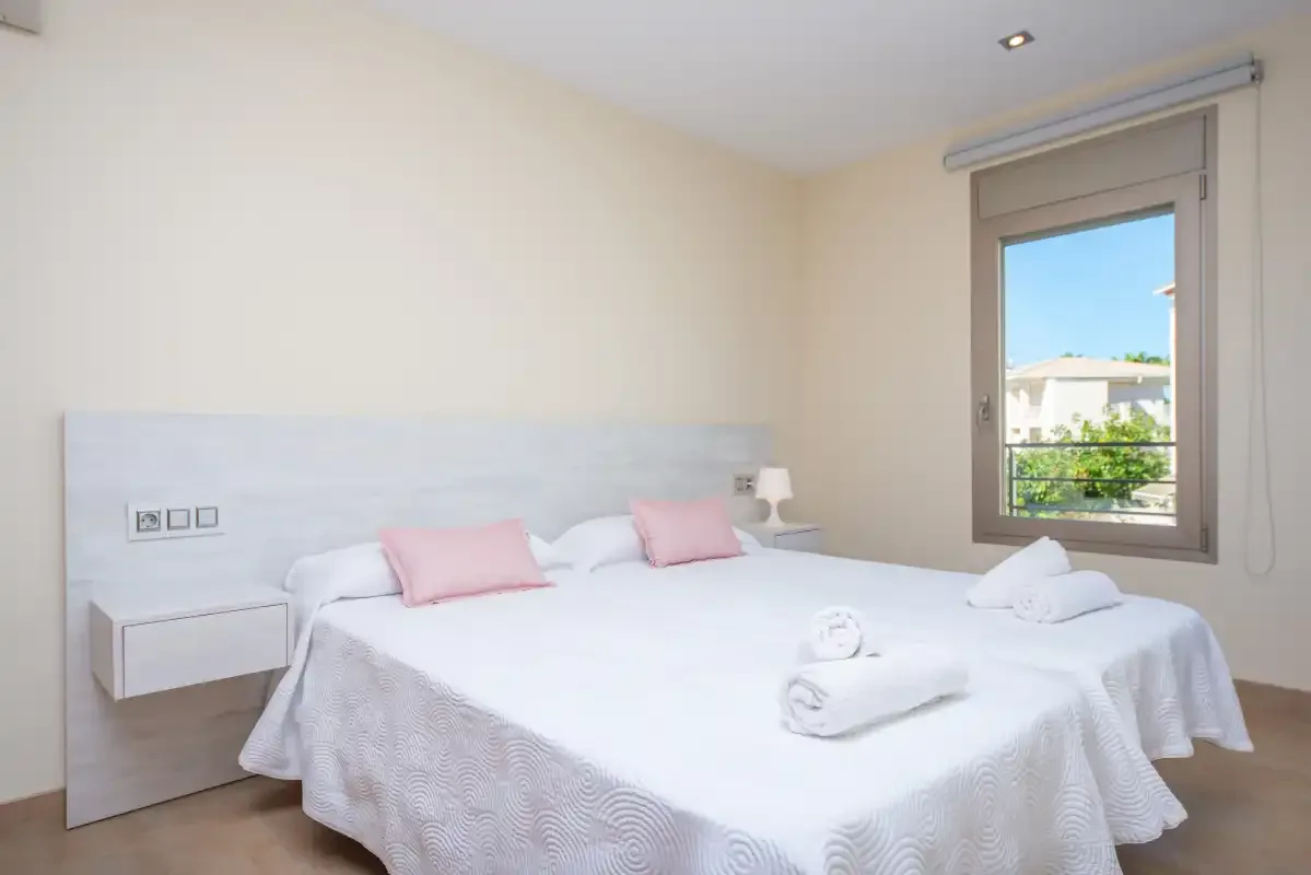 A bright bedroom with a neatly made bed and a window overlooking nearby buildings.