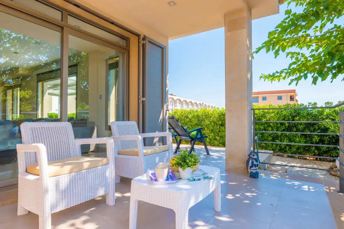A shaded patio has outdoor seating, a small table, and glass doors overlooking greenery.
