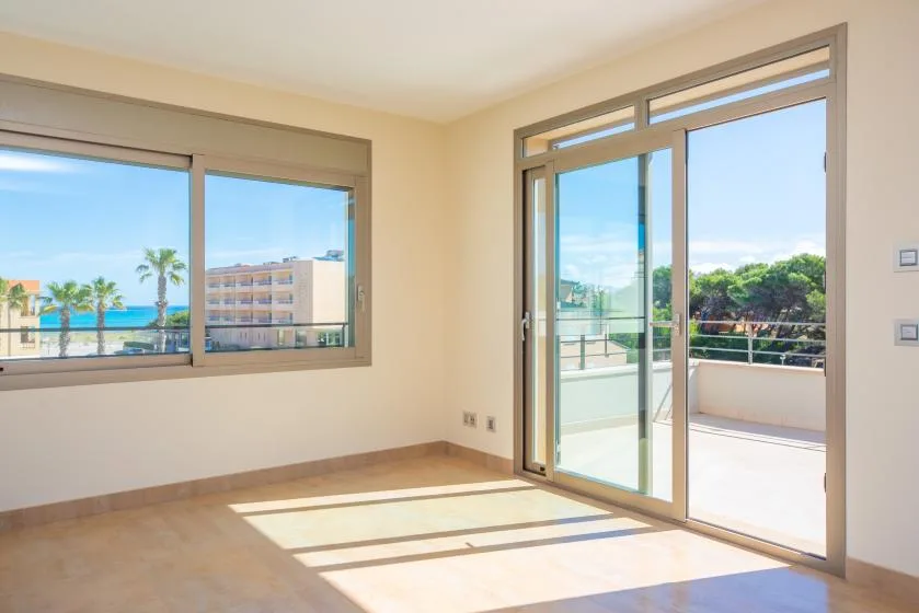 A bright, empty room opens onto a sunlit balcony through glass doors.