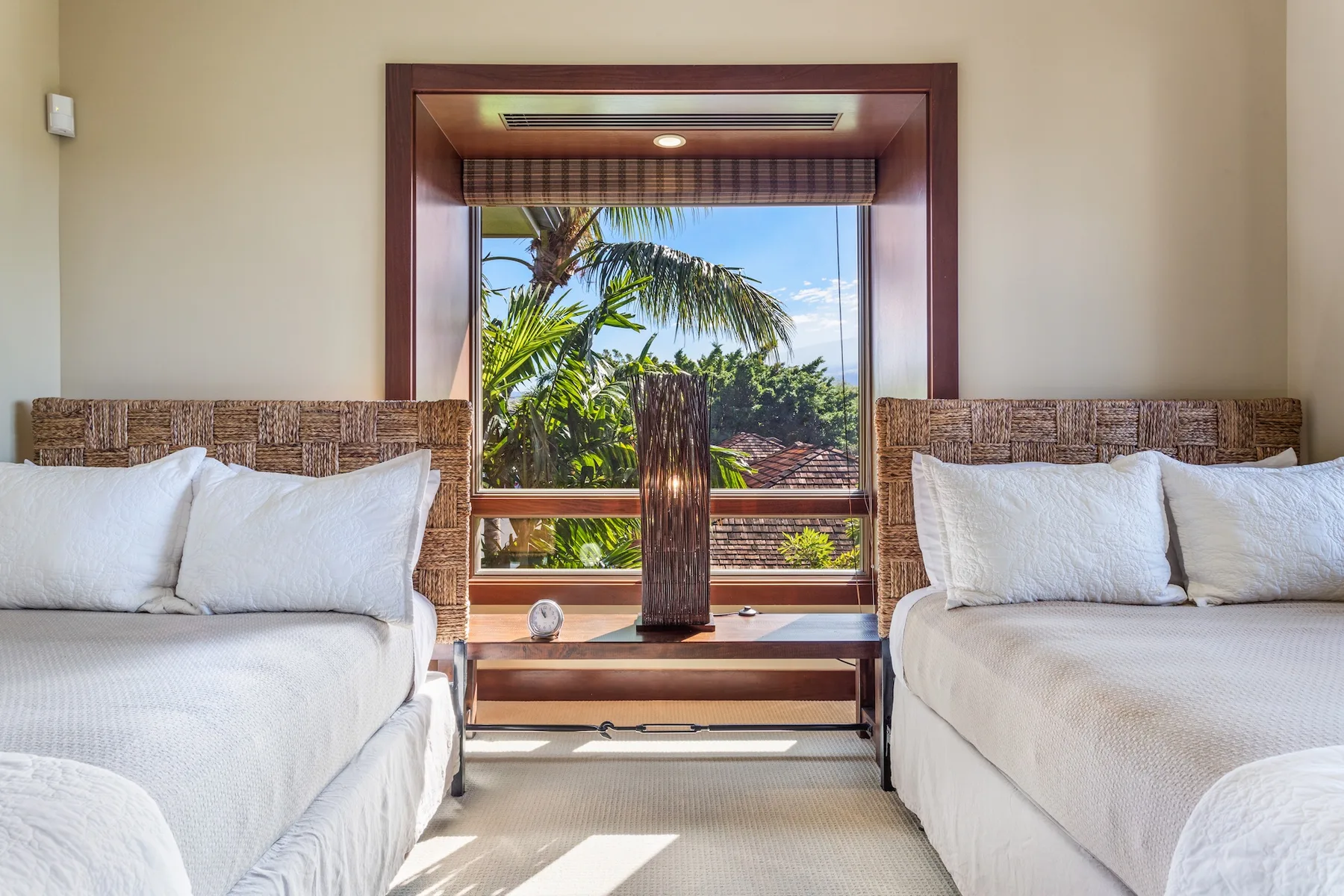 Un dormitorio luminoso con dos camas frente a una gran ventana con palmeras.