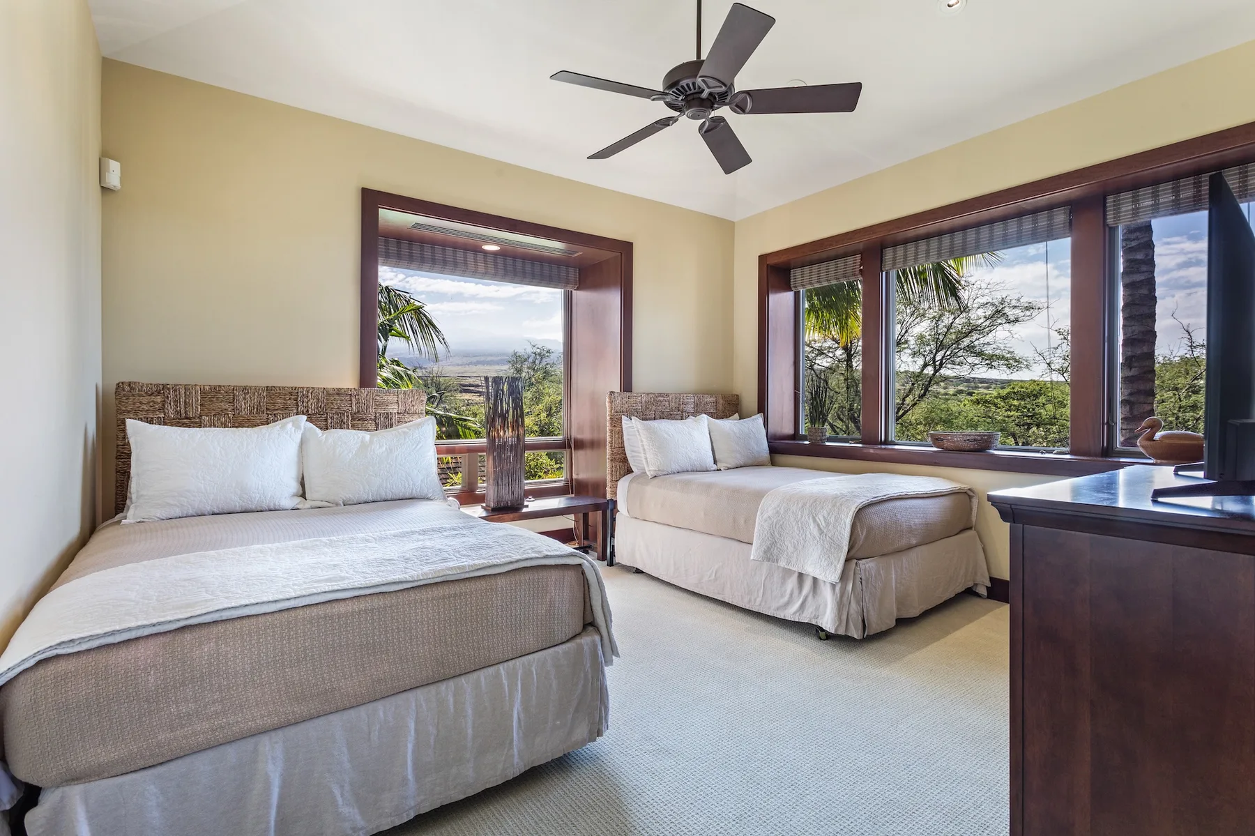 Un dormitorio luminoso con dos camas junto a grandes ventanas con vistas a la vegetación.