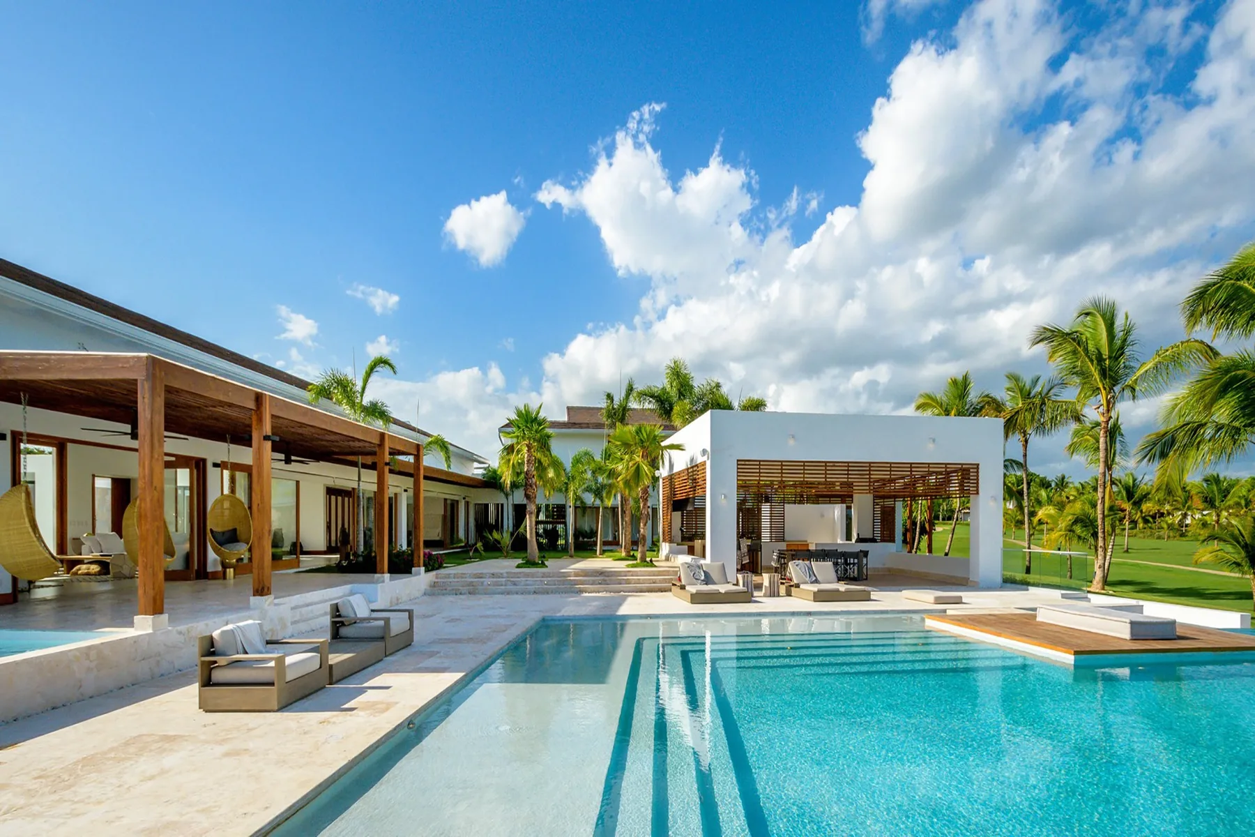 A pool beside a modern open-sided pavilion, with palm trees and outdoor seating.