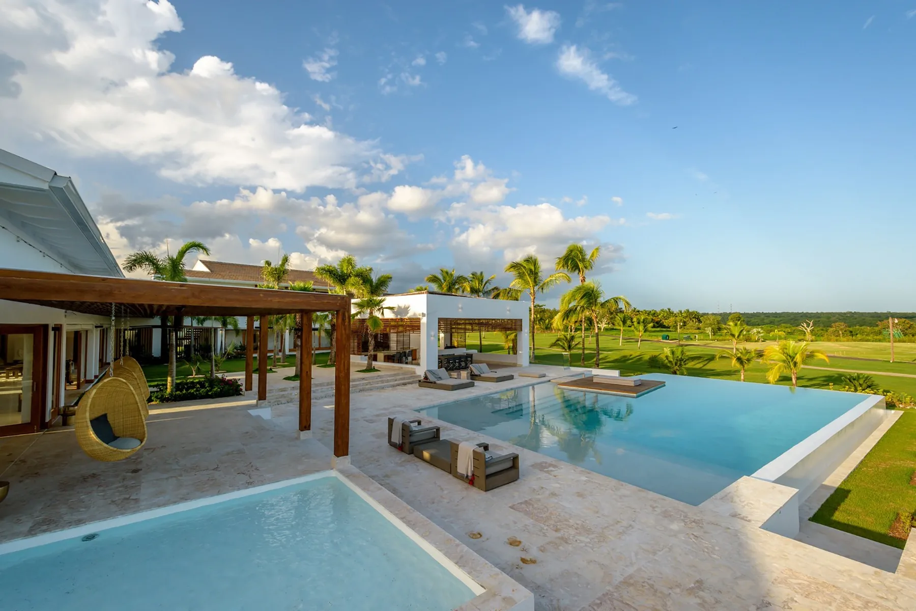 Una zona de piscina exterior junto a un patio cubierto, con vegetación tropical al fondo.