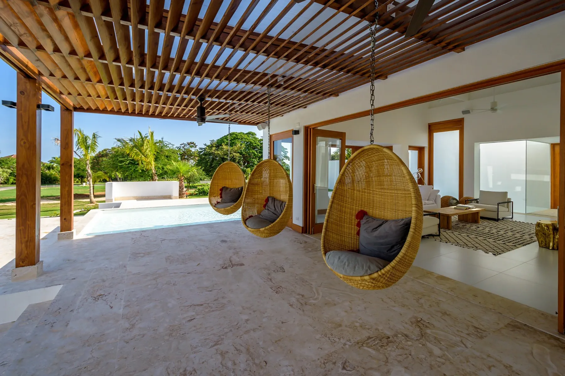 Un patio cubierto con asientos colgantes da a una piscina y vegetación.