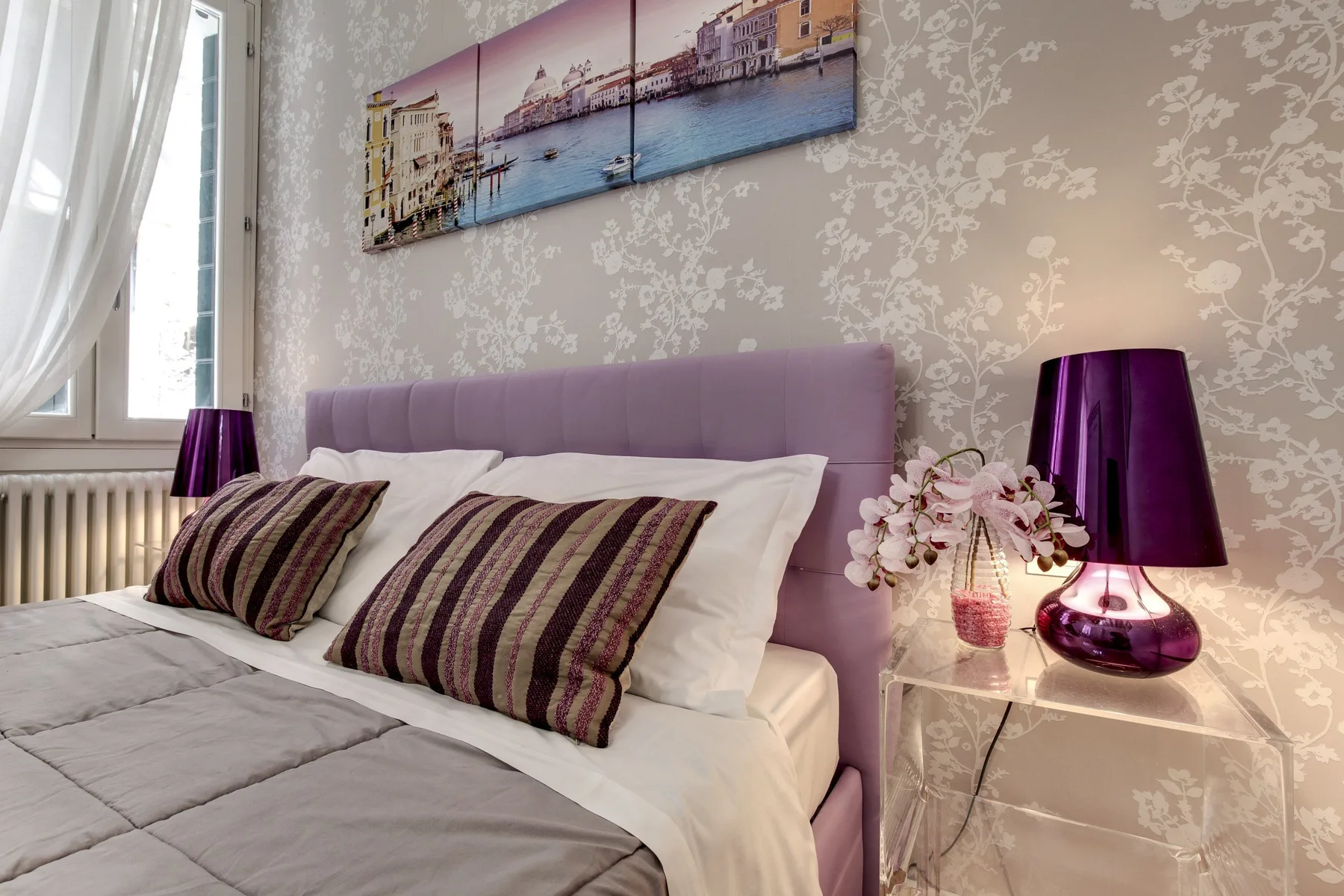 Un dormitorio con una cama, almohadas estampadas y lámparas moradas de noche.