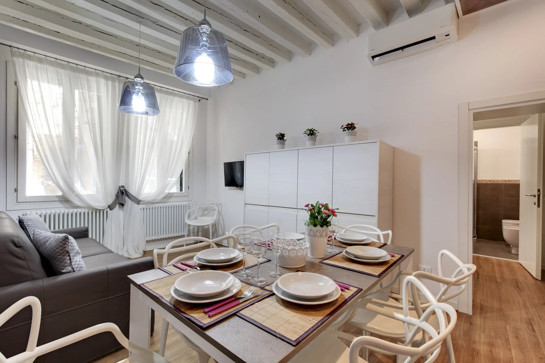 Comedor con una mesa puesta junto a un sofá y una ventana luminosa con cortinas.