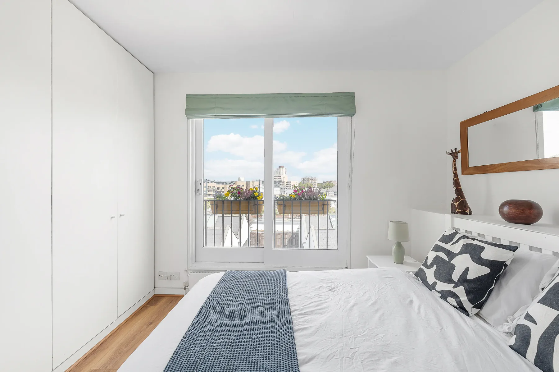 A bright bedroom with a bed beside a large window overlooking the city.