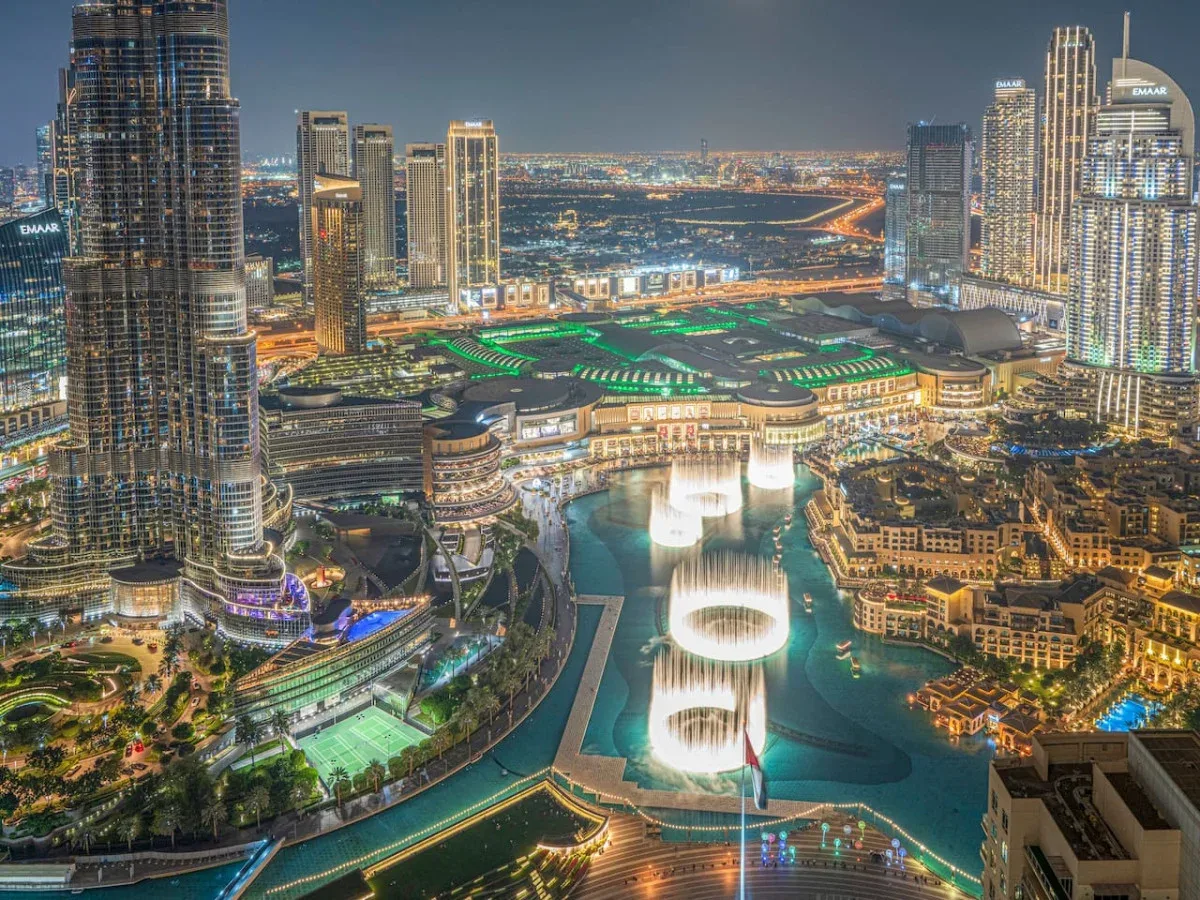 A nighttime cityscape shows illuminated towers around a large fountain and pool.