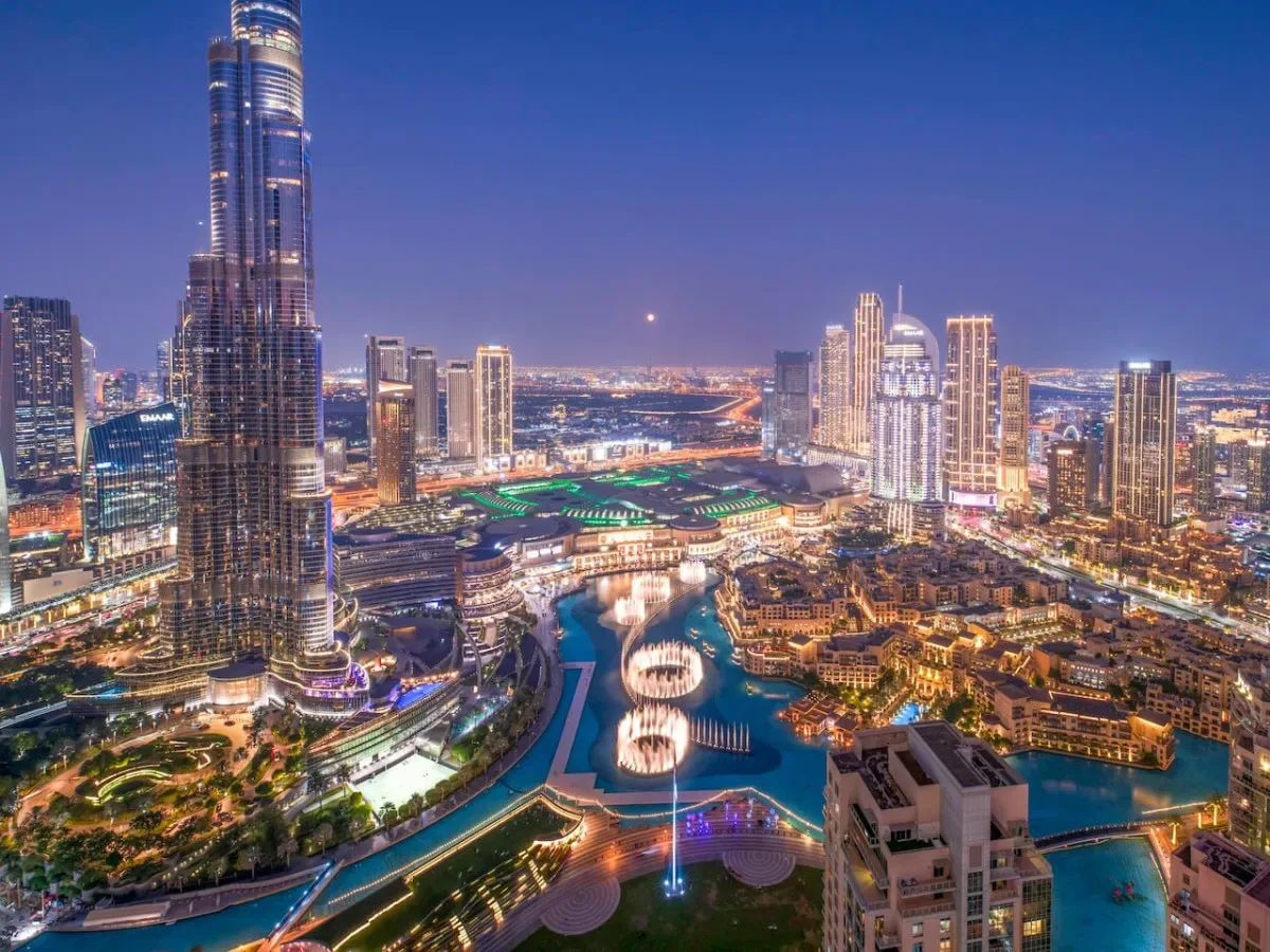 A nighttime city skyline surrounds a brightly illuminated waterfront and fountains.