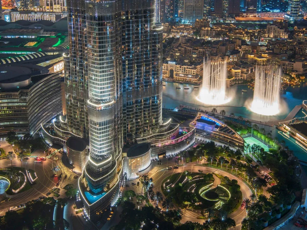 A nighttime cityscape features illuminated skyscrapers and fountains beside a landscaped plaza.