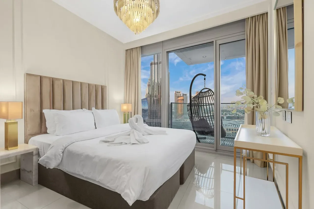 A bright bedroom features a neatly made bed beside large windows overlooking the city.