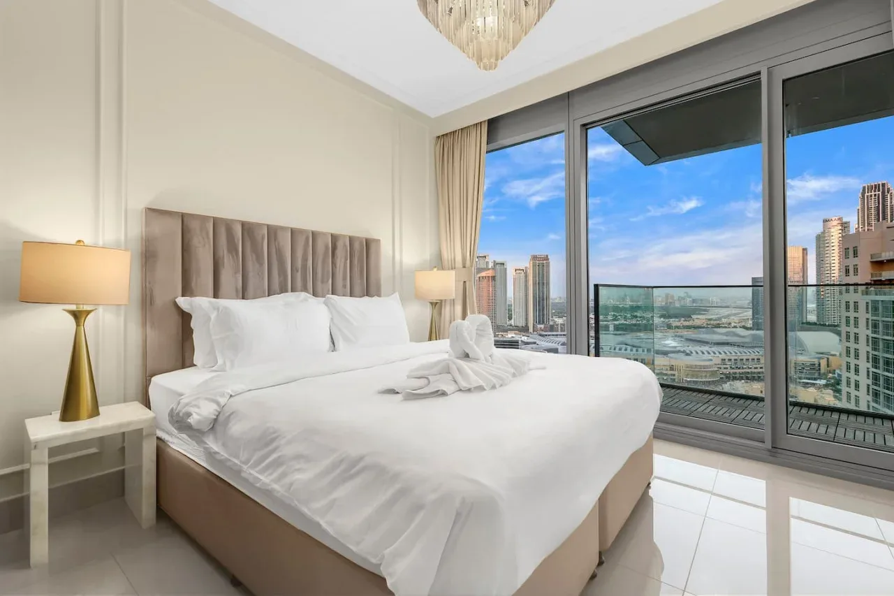 A bright bedroom with a large bed beside floor-to-ceiling windows overlooking city buildings.