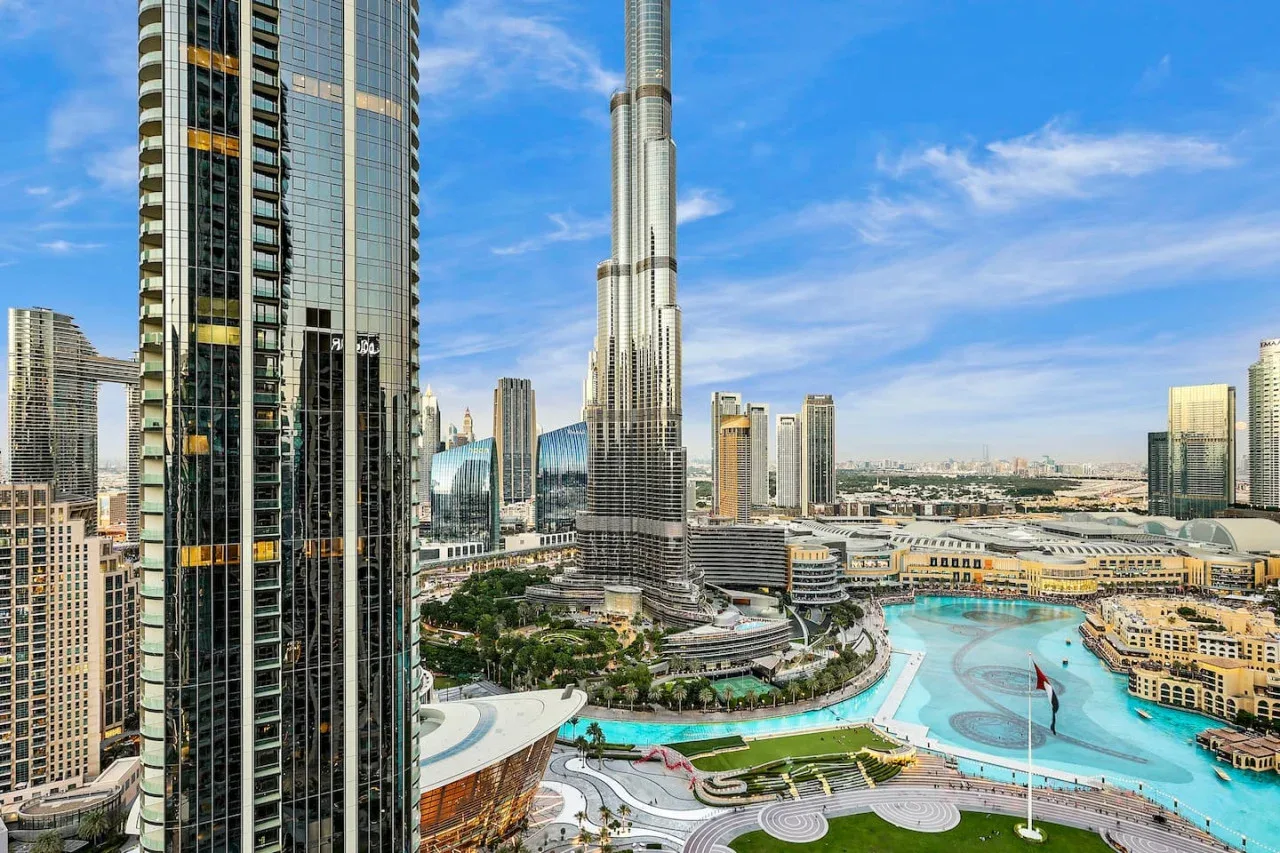 A city skyline overlooks a turquoise pool and landscaped grounds beneath a clear blue sky.
