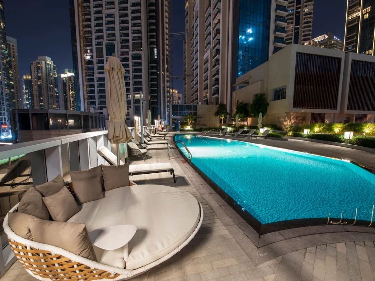 A rooftop pool at night is bordered by lounge seating and tall city buildings.